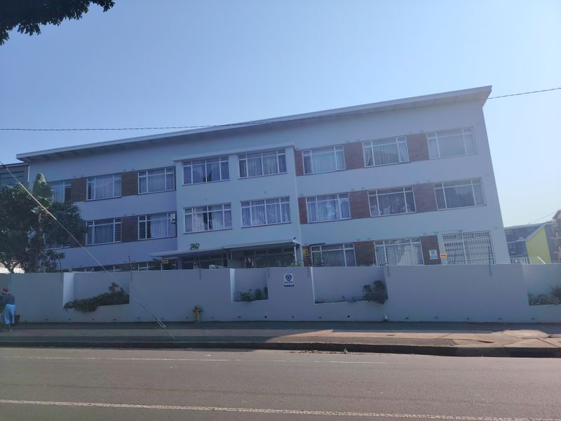 Street-facing view of a multi-story apartment building featuring a perimeter wall with electric fencing and classic casement window configurations.