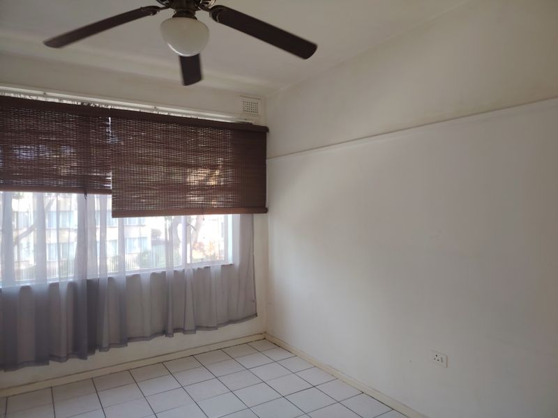 An empty bedroom featuring tiled flooring, a ceiling fan, and a large window with blinds, offering a blank canvas for potential renovation.