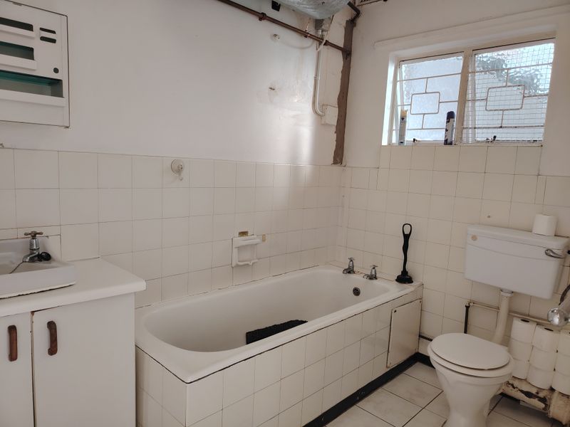 A dated bathroom featuring a tiled bathtub, wall-mounted basin, and toilet, with visible exposed plumbing and wall surface wear requiring cosmetic updates.