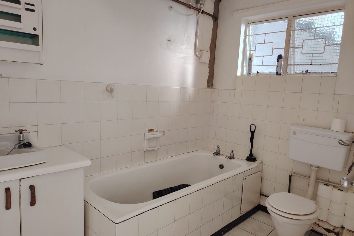 A dated bathroom featuring a tiled bathtub, wall-mounted basin, and toilet, with visible exposed plumbing and wall surface wear requiring cosmetic updates.