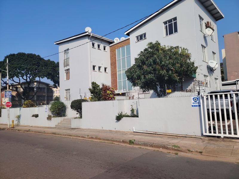 Front exterior view of a multi-story apartment building featuring a perimeter wall, electric fencing, and secure gated access from the street.