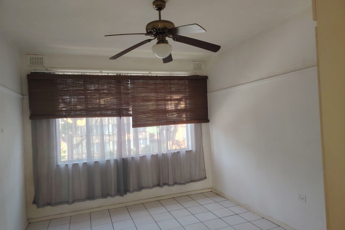 An empty living room featuring tiled flooring, a ceiling fan, and large windows with fitted blinds.