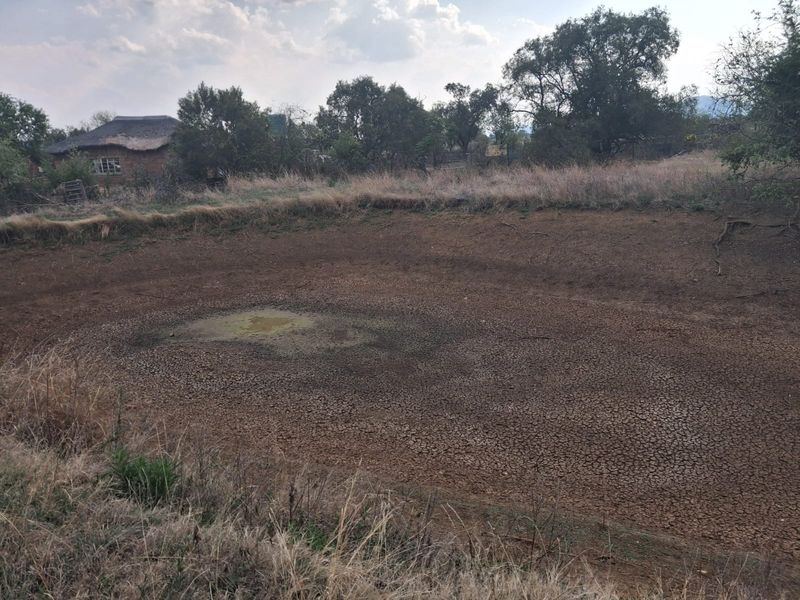 An exterior view of a dried-out earthen dam on the small holding property, showing significant lack of water and cracked ground.
