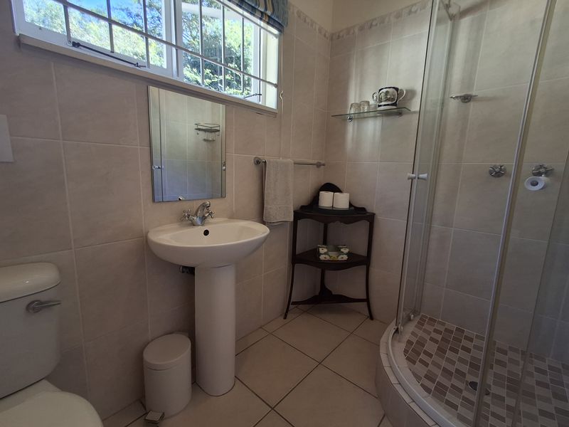 A functional bathroom featuring a pedestal basin, a corner shower enclosure, and neutral wall and floor tiling with natural light from a window.