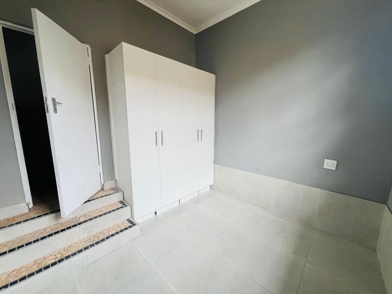 A clean, empty bedroom featuring neutral grey walls, white built-in cupboards, and tiled flooring with a small set of stairs leading to the entrance.