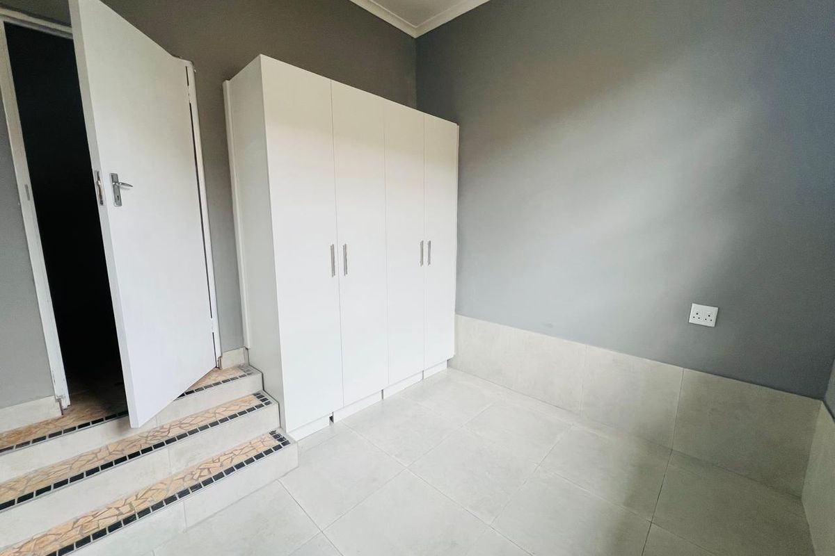 A clean, empty bedroom featuring neutral grey walls, white built-in cupboards, and tiled flooring with a small set of stairs leading to the entrance.