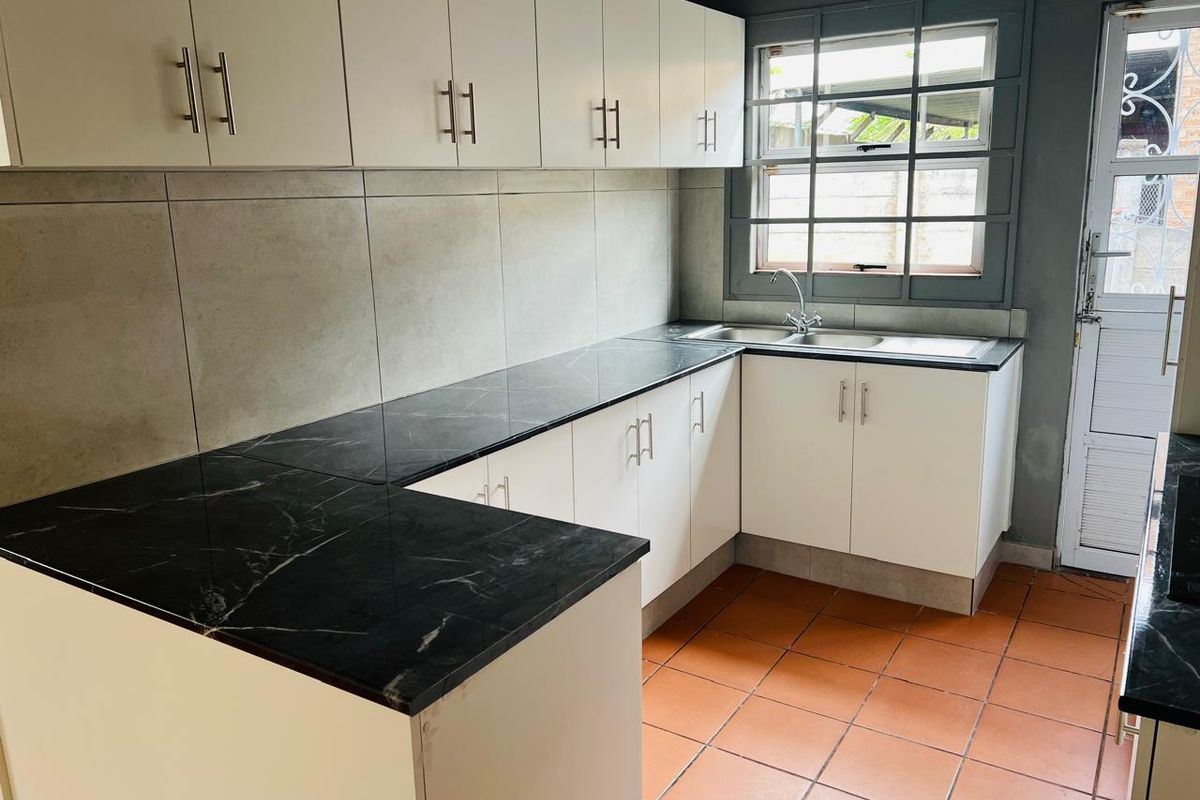 A clean, functional kitchen featuring white cabinetry, black marble-effect countertops, terracotta flooring, and a large window providing natural light.