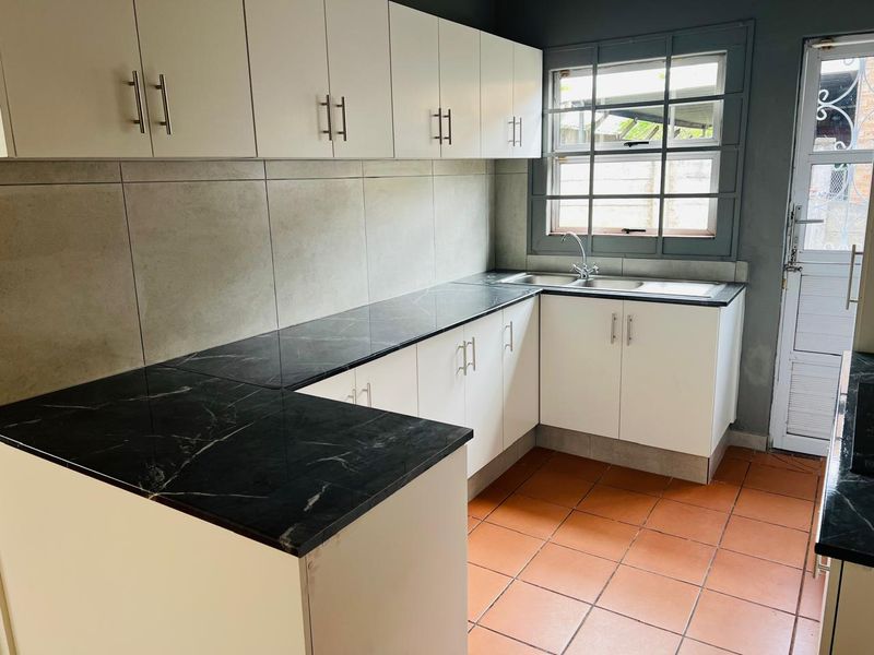 A clean, functional kitchen featuring white cabinetry, black marble-effect countertops, terracotta flooring, and a large window providing natural light.