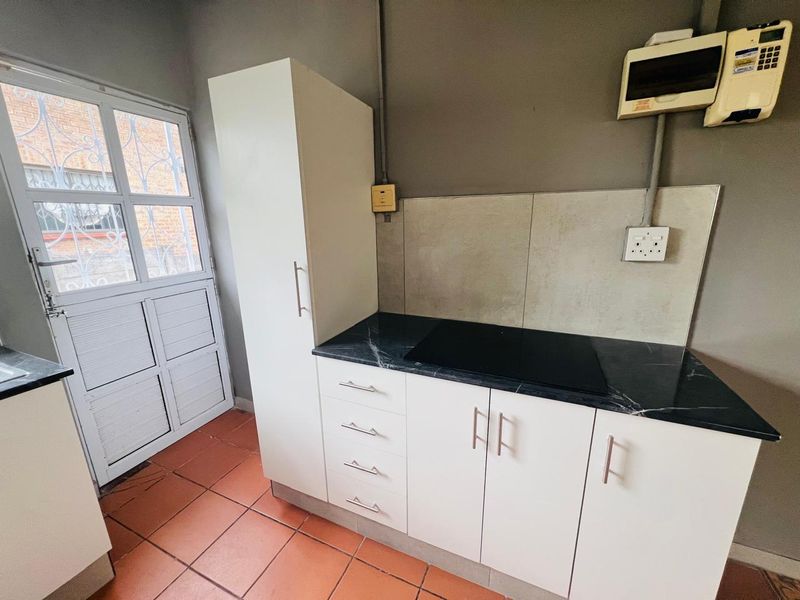 A compact kitchen area featuring white cabinetry, dark laminate countertops, an integrated electric hob, and terracotta-style floor tiles with direct exterior access.