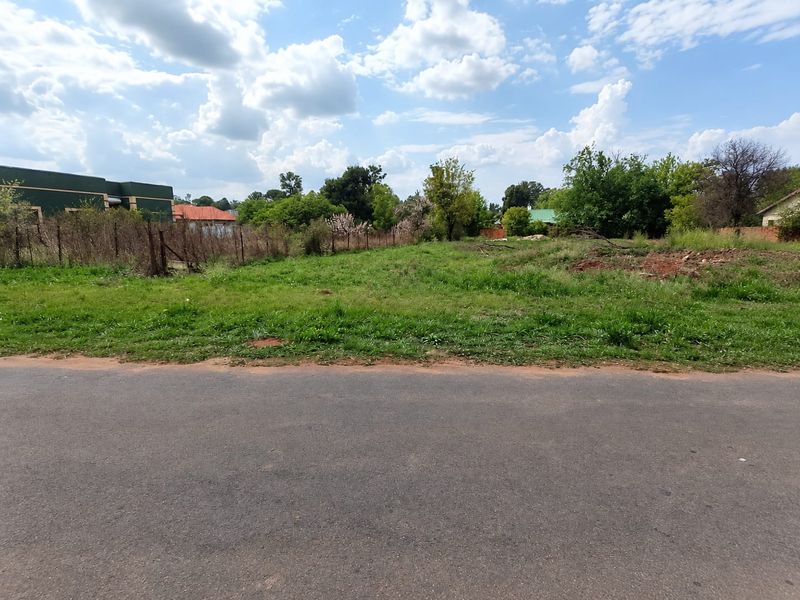 A vacant residential plot of land featuring grass cover, bordered by wire fencing and accessible via an adjacent asphalt road.