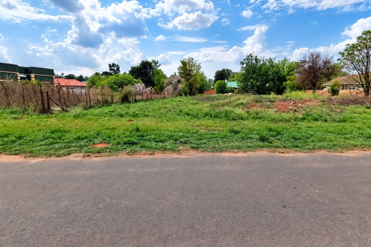 A street-level view of a vacant residential plot featuring grass coverage and perimeter wire fencing, situated along an established asphalt road.