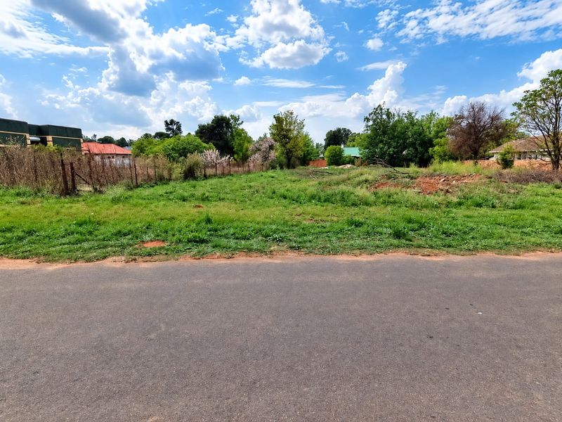 A street-level view of a vacant residential plot featuring grass coverage and perimeter wire fencing, situated along an established asphalt road.