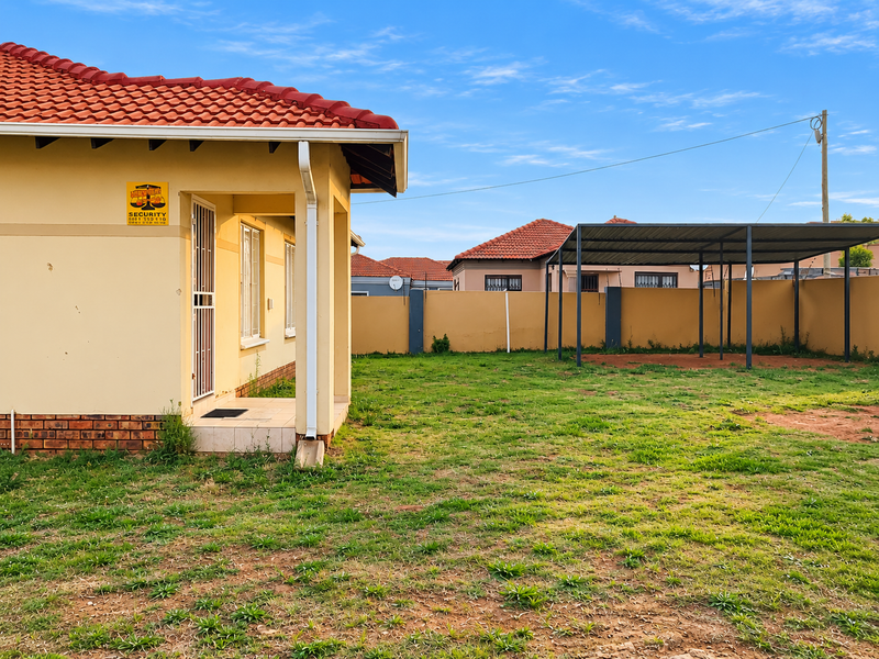 Rear view of the property featuring a tiled roof, a covered carport structure, and a spacious backyard enclosed by a perimeter wall.
