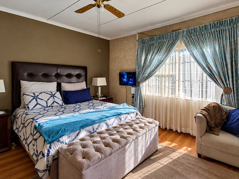 A well-lit bedroom featuring hardwood flooring, a ceiling fan, and large windows with security bars, offering a comfortable and functional living space.