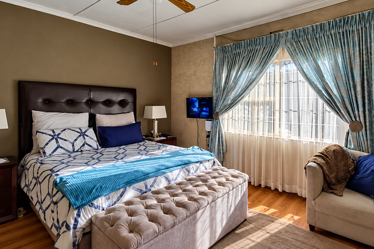 A well-lit bedroom featuring hardwood flooring, a ceiling fan, and large windows with security bars, offering a comfortable and functional living space.