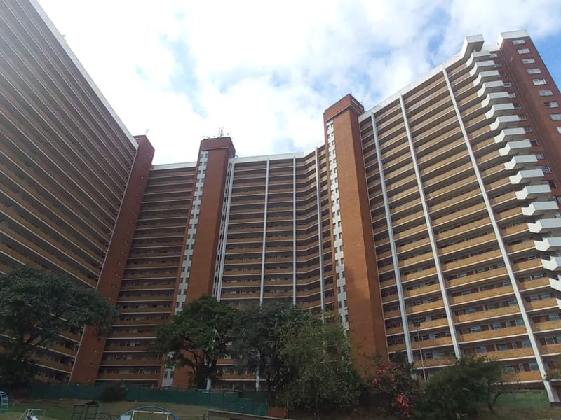 A low-angle view of a large, multi-story brick residential apartment building featuring numerous balconies and established surrounding greenery.