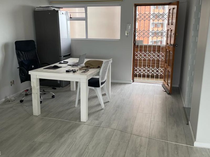 A bright living area featuring modern light-toned laminate flooring, a secure entrance with a metal gate, and natural light from the window.