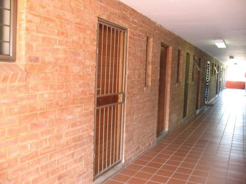 A long, open-air apartment hallway featuring exposed brick walls, tiled flooring, and secure metal gates leading to individual units.