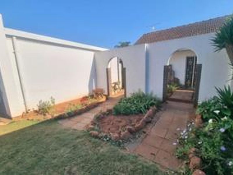 A rear exterior view featuring white rendered walls, arched entryways, paved walkways, and established garden beds.
