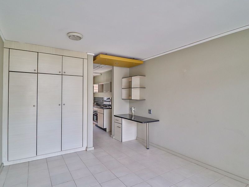 Open plan living area featuring built-in storage cupboards, a fixed desk workspace, and tiled flooring, with a view into the adjacent kitchen area.