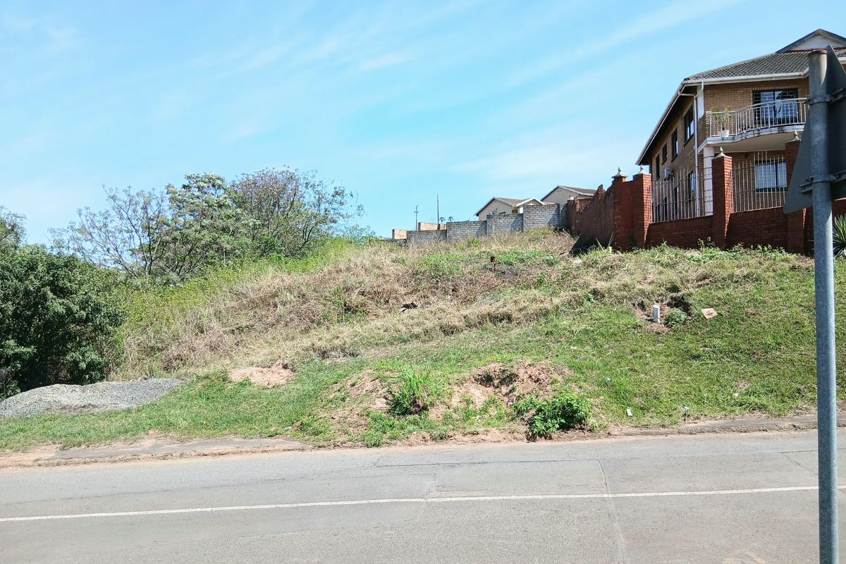 A vacant residential plot featuring a sloped landscape, adjacent to an existing brick boundary wall and paved street.