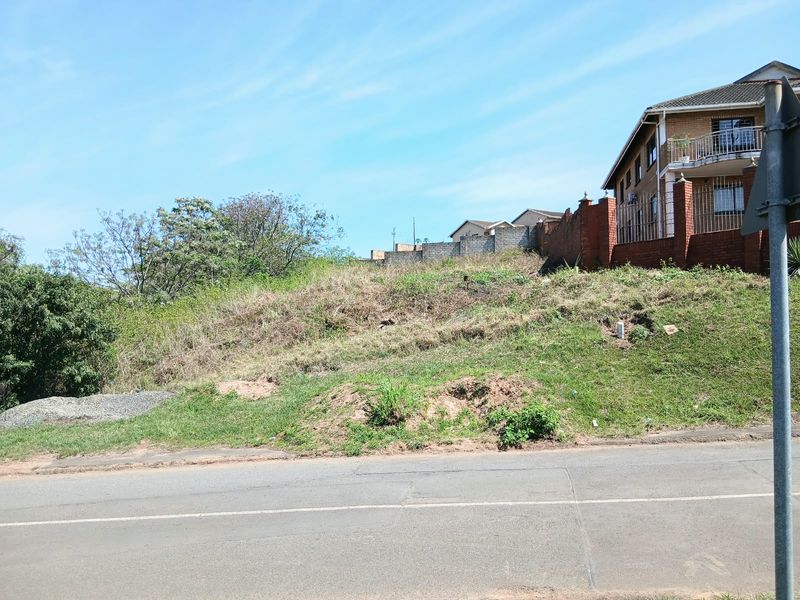 A vacant residential plot featuring a sloped landscape, adjacent to an existing brick boundary wall and paved street.