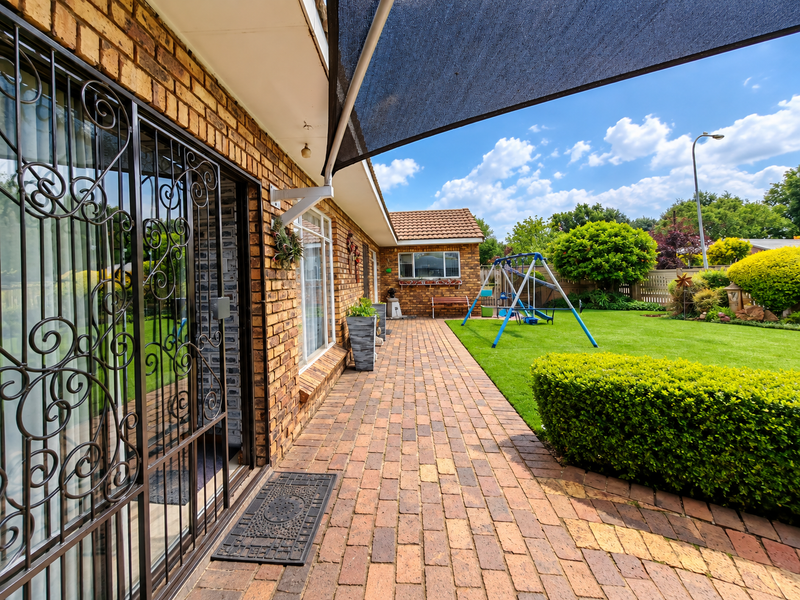 A well-maintained paved patio area featuring face brick walls, a security gate, and a shade sail, overlooking a landscaped garden.
