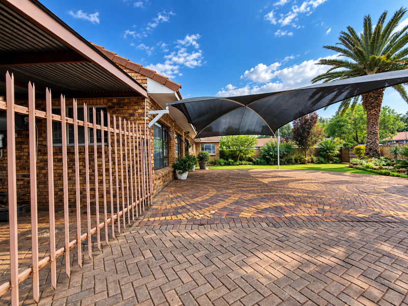 Rear exterior view featuring extensive brick paving, a large shade sail, covered patio area, and secure metal fencing.
