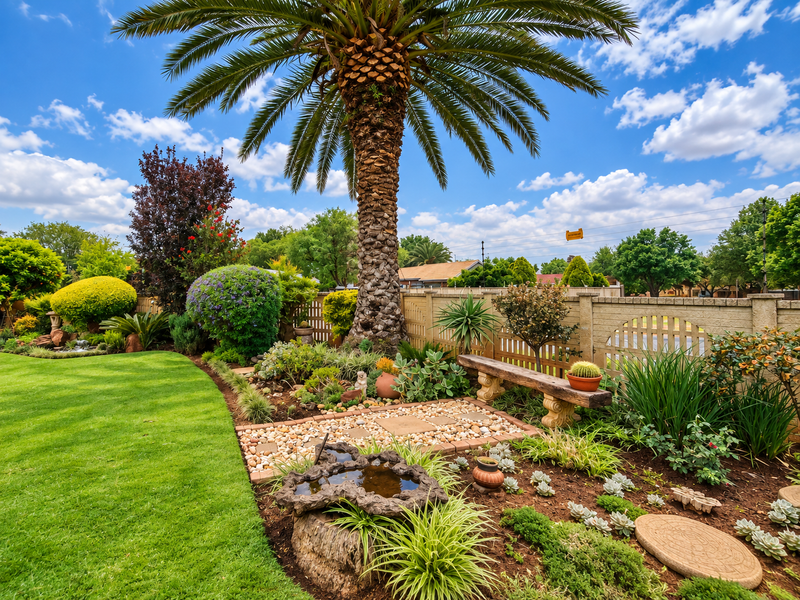 A beautifully landscaped garden featuring a mature palm tree, manicured lawn, stone pathways, and a decorative precast concrete boundary wall.