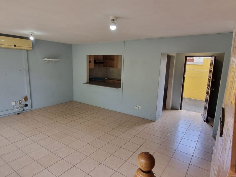 An open-plan living area featuring tiled flooring, an air conditioning unit, and a pass-through window connecting to the kitchen, with an adjacent staircase.