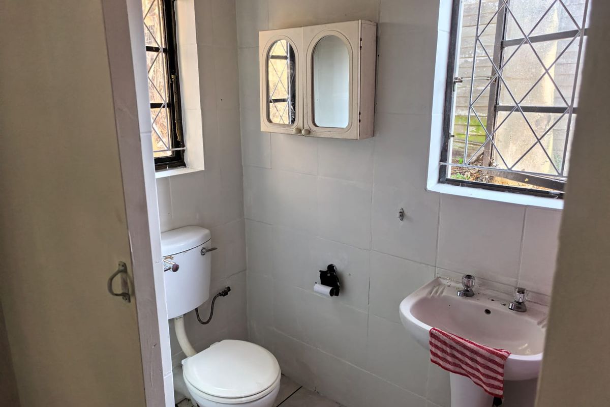 A compact, dated bathroom featuring a pedestal basin, toilet, and wall-mounted medicine cabinet, with original leaded glass windows and tiled walls.