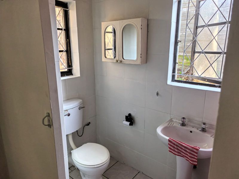 A compact, dated bathroom featuring a pedestal basin, toilet, and wall-mounted medicine cabinet, with original leaded glass windows and tiled walls.