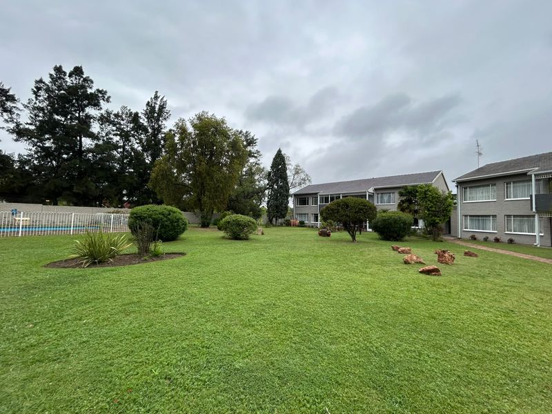 A spacious communal garden area featuring a well-maintained lawn, mature trees, a swimming pool, and paved walkways between the residential buildings.