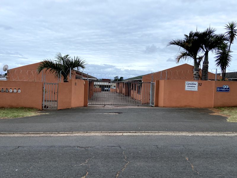 Front entrance view of Caroline Gardens townhouse complex, featuring a gated security entrance, perimeter wall with razor wire, and paved driveway access.