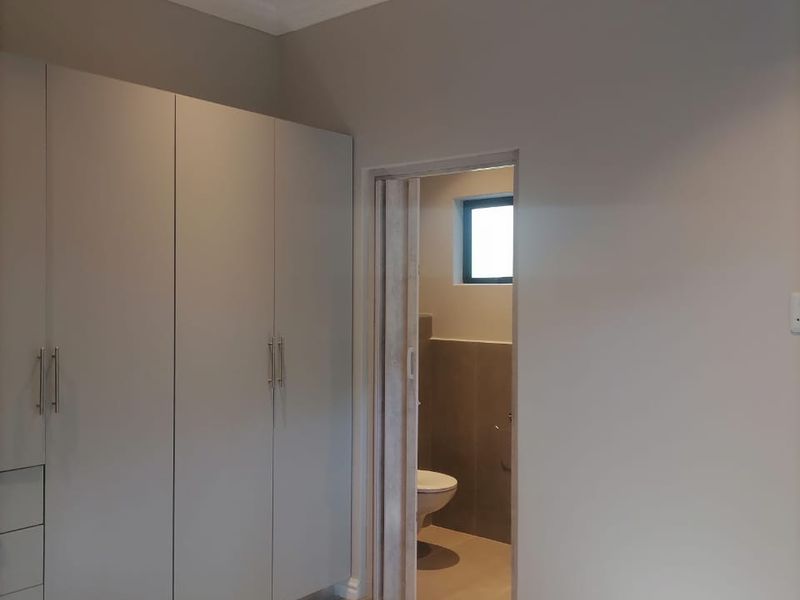 A clean, modern bedroom featuring neutral walls, tiled flooring, and extensive built-in cupboards, with an adjoining en-suite bathroom visible through the doorway.