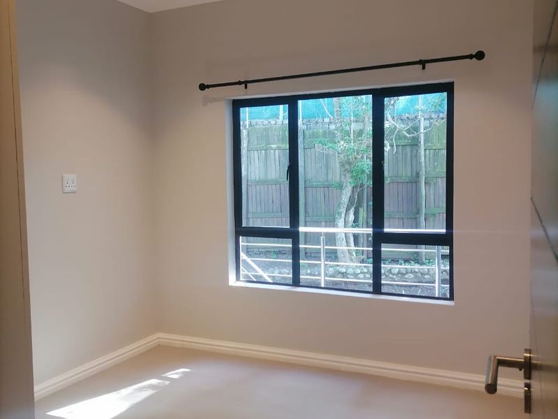 A clean, empty bedroom featuring neutral walls, recessed downlighting, and a large window with a black aluminium frame.