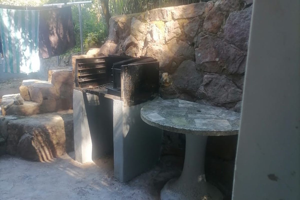 Outdoor rear patio area featuring a built-in braai, stone retaining wall, and a fixed concrete pedestal table in a garden setting.