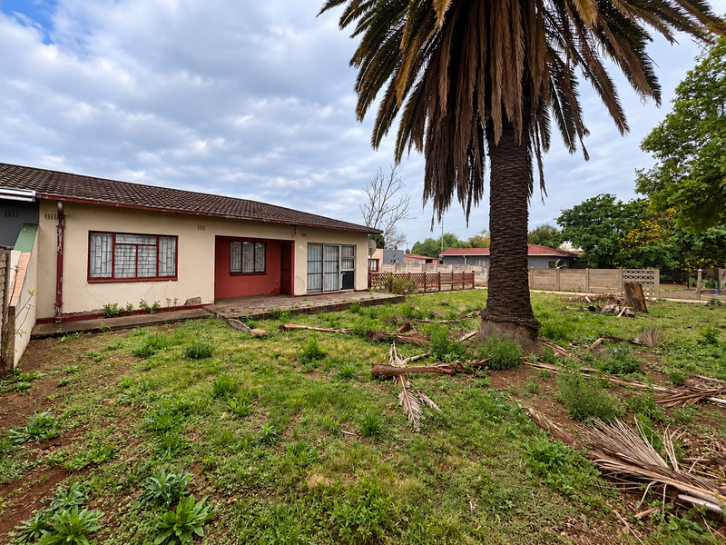 Rear view of a single-story house featuring a stone patio, sliding glass doors, and a large garden area requiring landscaping maintenance.