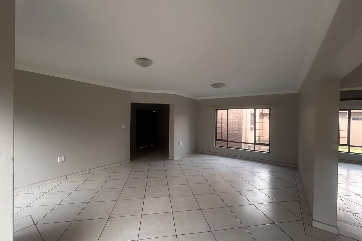 A spacious, empty living area featuring neutral wall tones, ceramic floor tiles, and large windows with security bars, offering a clean, blank canvas.