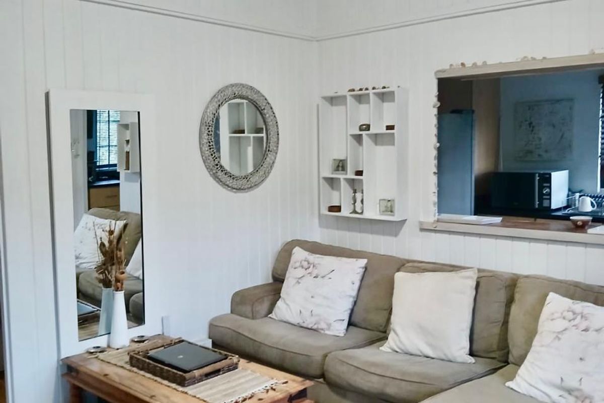 A cozy living room featuring timber flooring, white timber wall panelling, a ceiling fan, and a convenient serving hatch connecting to the kitchen area.