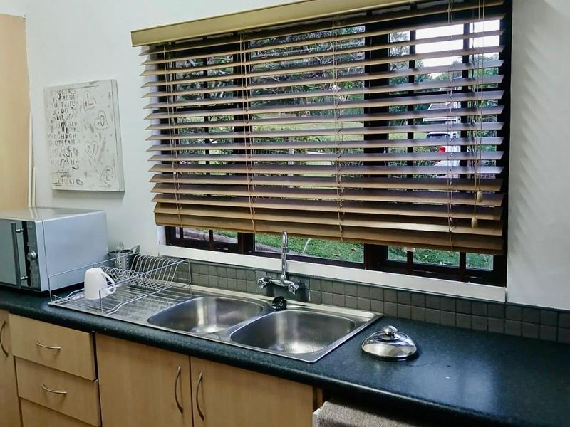 A functional kitchen area featuring a double bowl stainless steel sink, laminate cabinetry, and a tiled splashback with venetian blinds over the window.