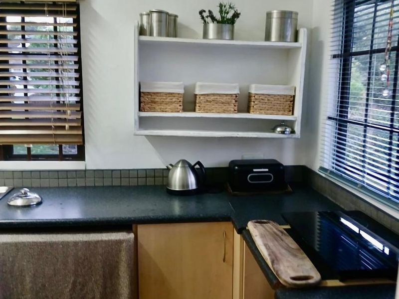 A compact kitchen area featuring dark laminate countertops, light wood-tone cabinetry, tiled splashbacks, and functional wall-mounted shelving under dual windows.