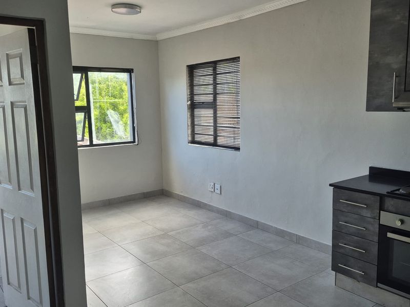 An open-plan living and kitchen area featuring modern grey cabinetry, built-in appliances, neutral tiled flooring, and large windows with venetian blinds.