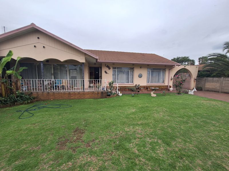 Rear view of a single-story house featuring a covered patio, tiled roof, and a spacious lawn with paved driveway and boundary wall.
