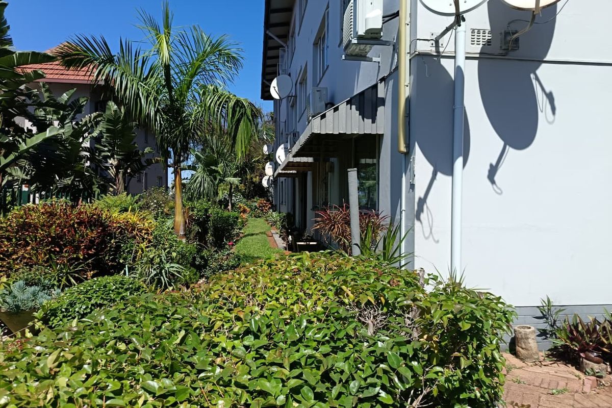 A view of the rear exterior of the flat building, featuring a landscaped garden area, paved pathways, and mounted satellite dishes.