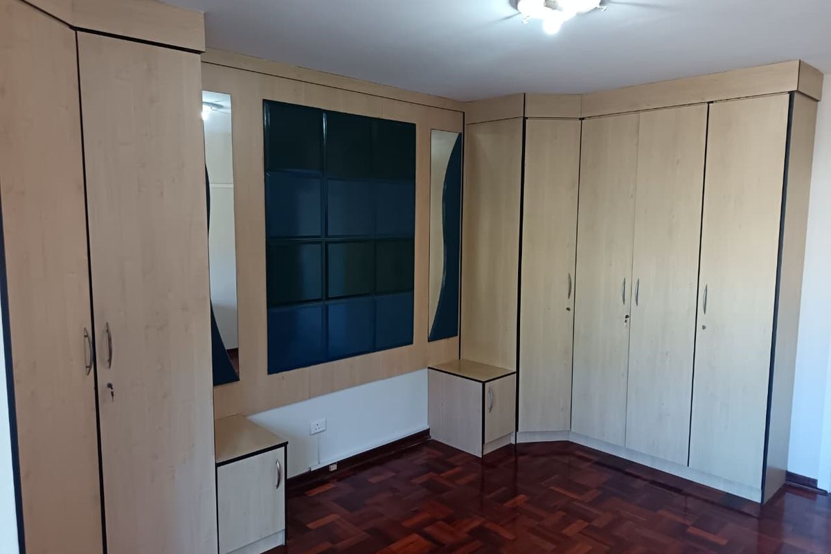 A spacious bedroom featuring extensive built-in wooden cupboards, integrated headboard with mirrored accents, and well-maintained parquet flooring.