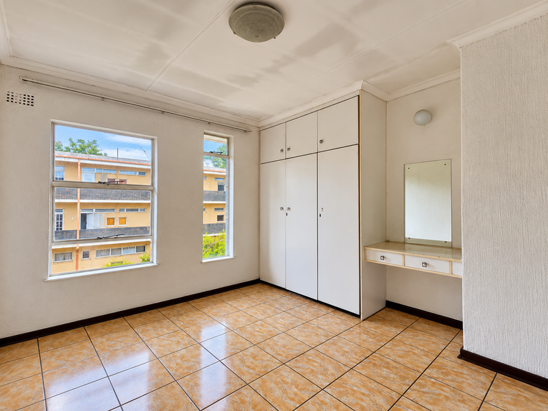 A bright, empty bedroom featuring built-in cupboards, a vanity area with a mirror, and tiled flooring, offering a blank canvas for personalization.