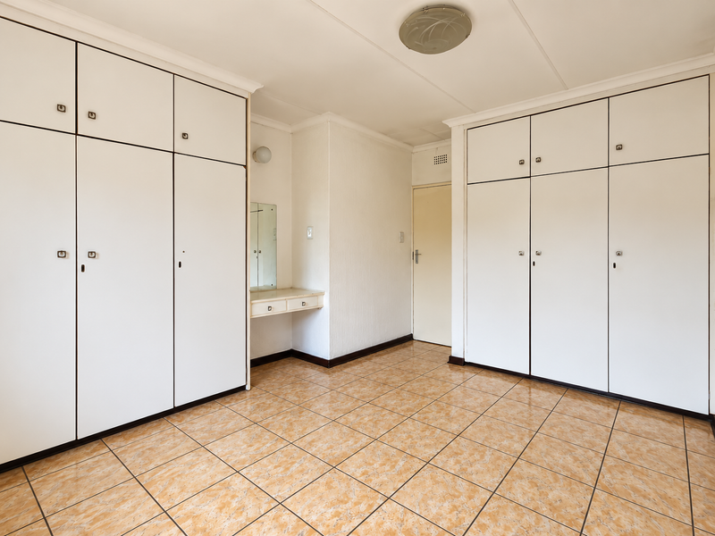 A spacious bedroom featuring extensive built-in cupboard storage, a small integrated vanity area, and durable tiled flooring throughout.