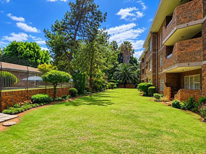 A well-maintained rear garden area featuring a lush lawn, landscaped borders, and a multi-story brick apartment building with private balconies.
