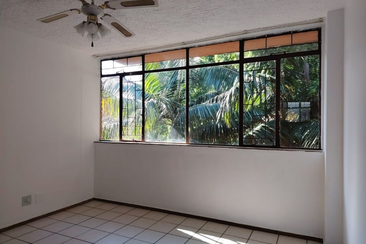 An empty bedroom featuring tiled flooring, a ceiling fan, and large metal-framed windows with a view of lush greenery.